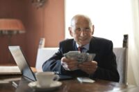 confident senior businessman holding money in hands while sitting at table near laptop