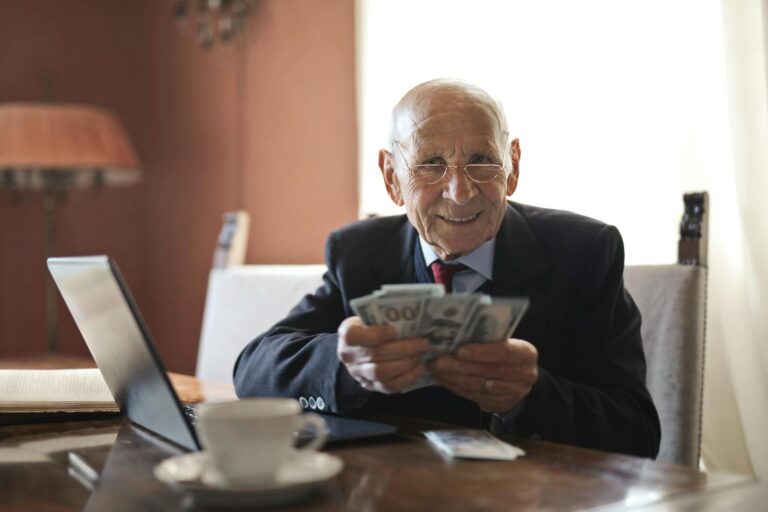 confident senior businessman holding money in hands while sitting at table near laptop